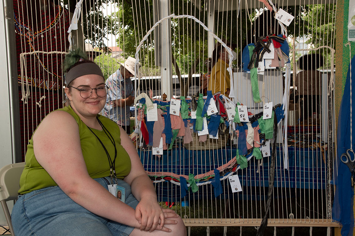 A person in green tank top and jean shorts sits in front of a vertical loom, several white parallel white strands, with scraps of pink, green, and blue fabric woven into it, the start of a spiraling shape.