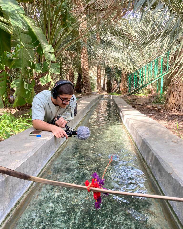 Mansour Al Heera, wearing gray shirt and headphones, holds an audio recorder over a cement-lined channel of water, running through an area of dense palm trees.