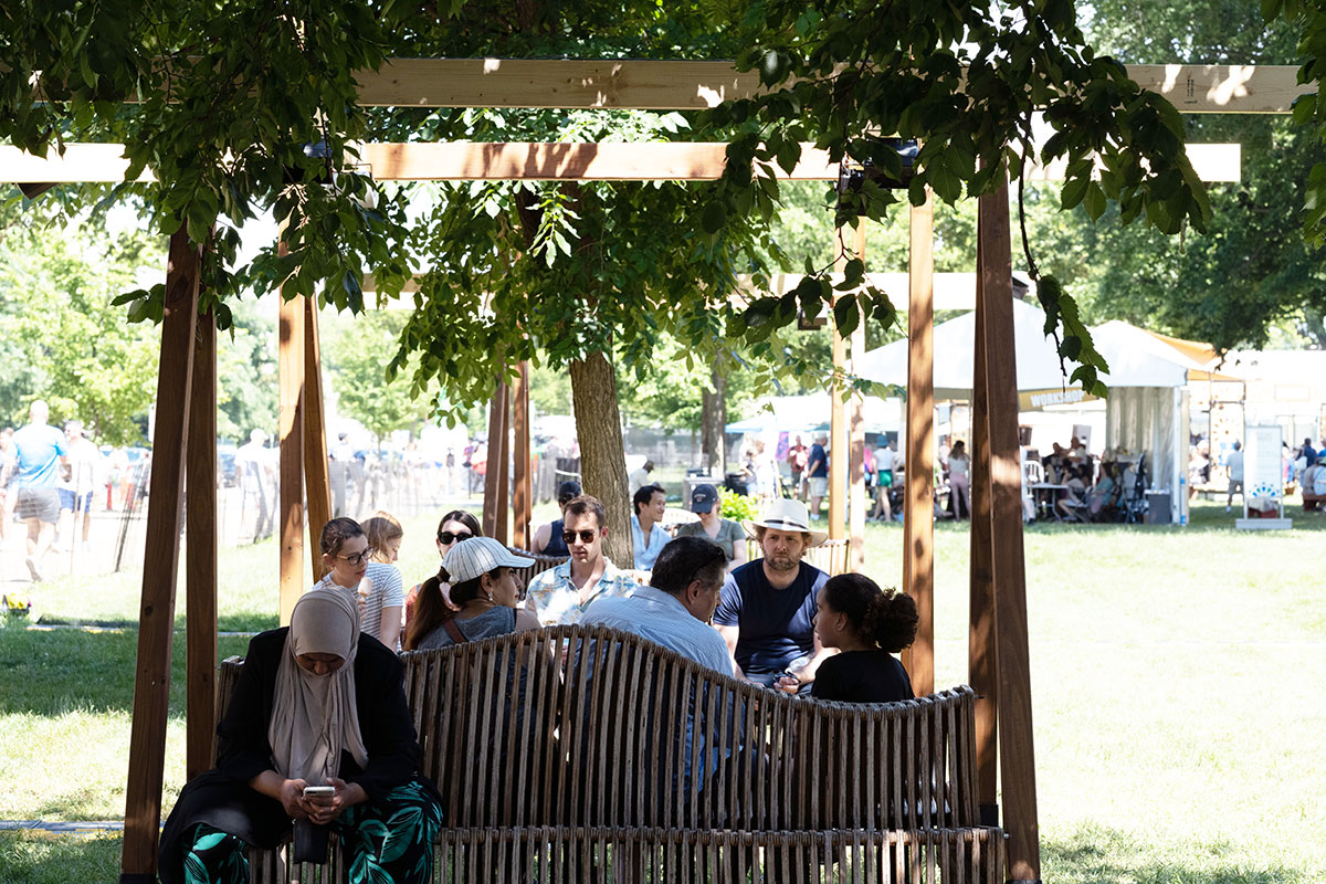 Several people sit on a wooden structure of slatted benches and beams overhead, in a grassy area under a tree.