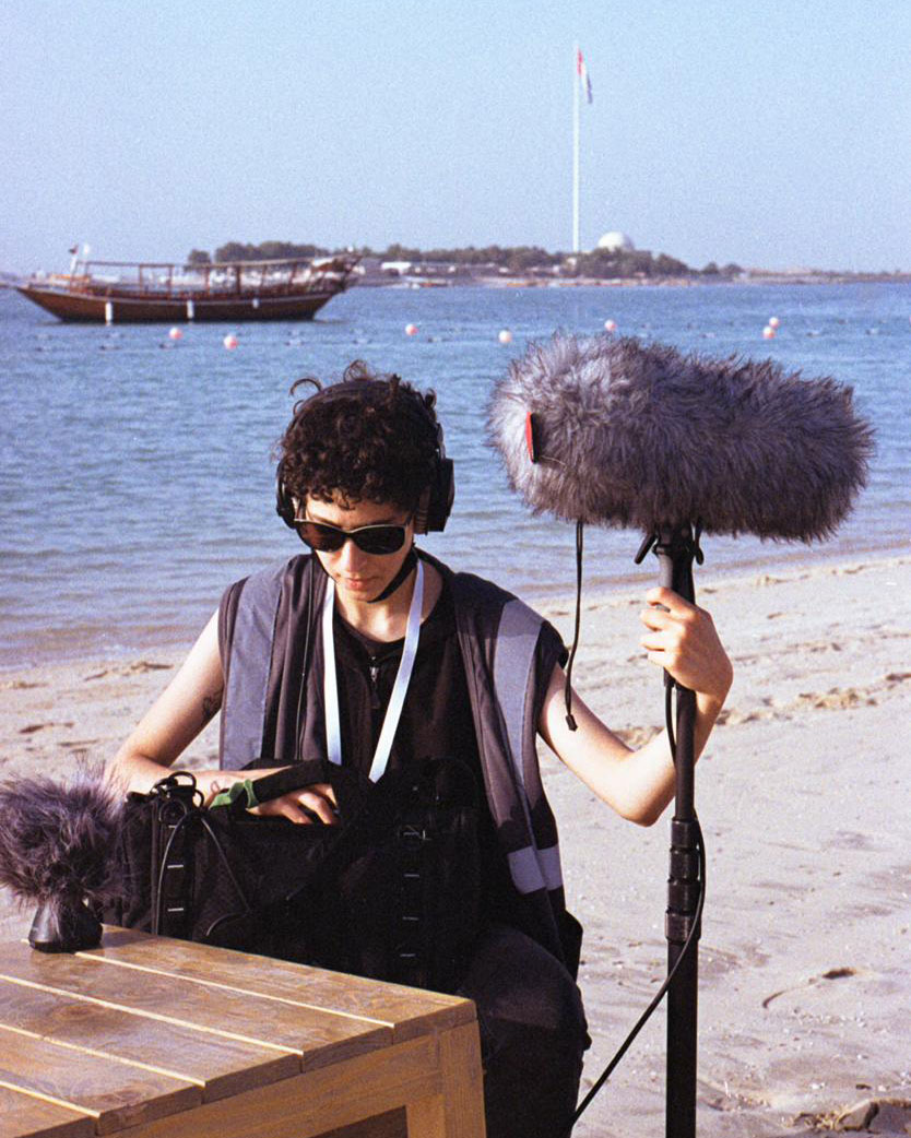 Safeya Al Blooshi, dressed in black, holds up a boom microphone with a fuzzy gray windscreen on a beach, along the water.