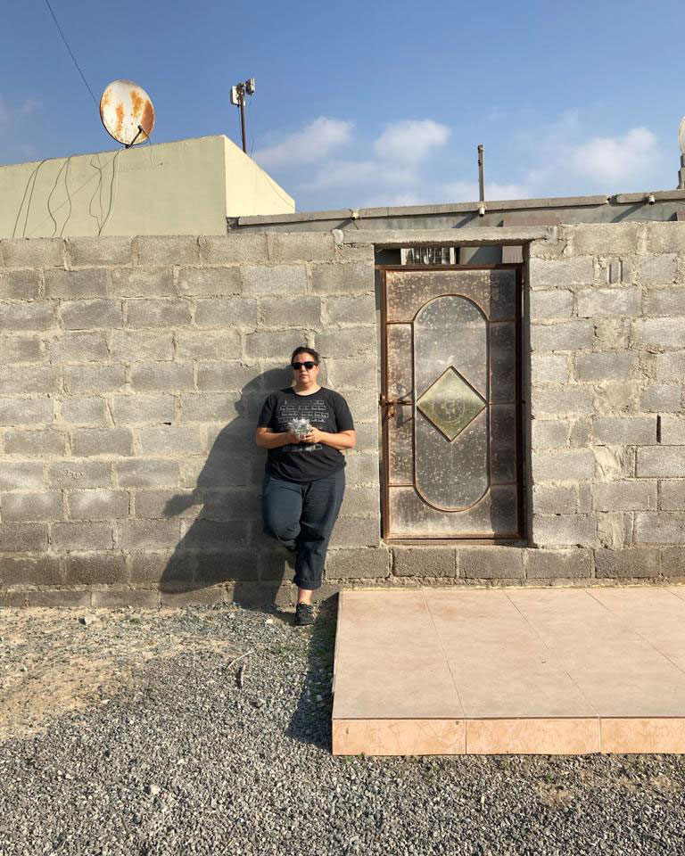 Diana Chester, wearing black shirt, pants, shoes, and sunglasses, leans against a gray brick wall outside, next to a doorway.