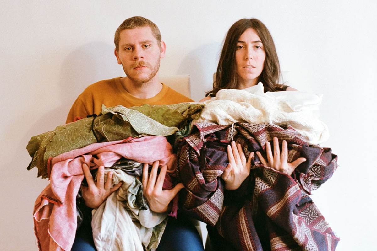 Two people pose, holding up piles of worn looking fabric. Neither is smiling.