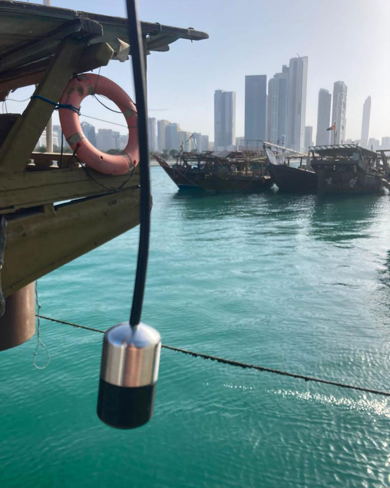 A black and silver cyclindrical microphone dangles by a cord over a wooden platform above turquoise water. Wooden boats and the skyscapers of Dubai loom in the background.