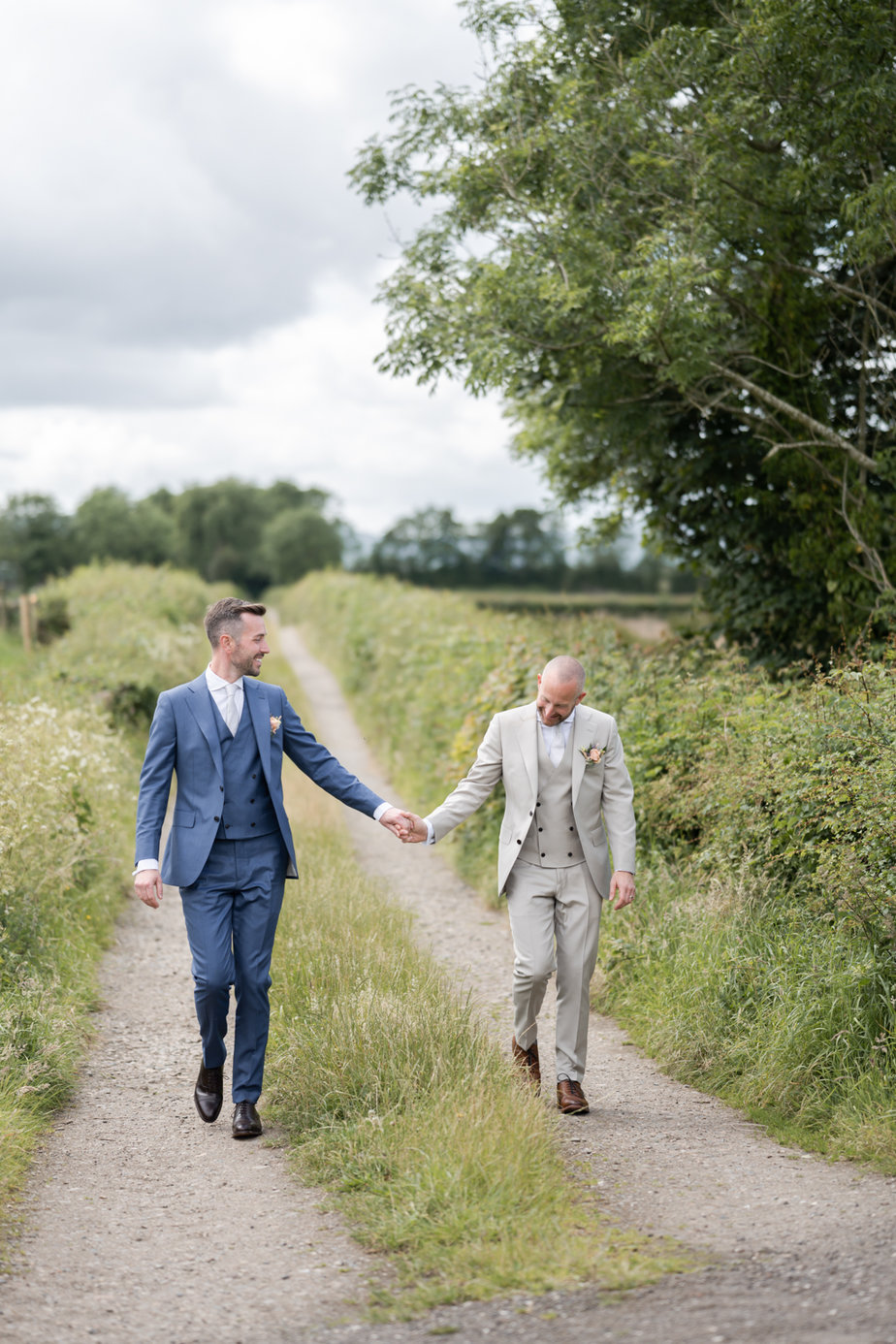 Grooms standing in field beside farm holding hands