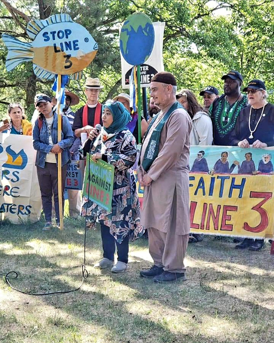 A man and woman, both wearing long tunics and head coverings, stand in front of a group of protestors outdoors, as the woman speaks into a microphone. Their protest signs include Muslims Against Line 3, Stop Line 3 on the image of a fish, and an iamge of a globe. 