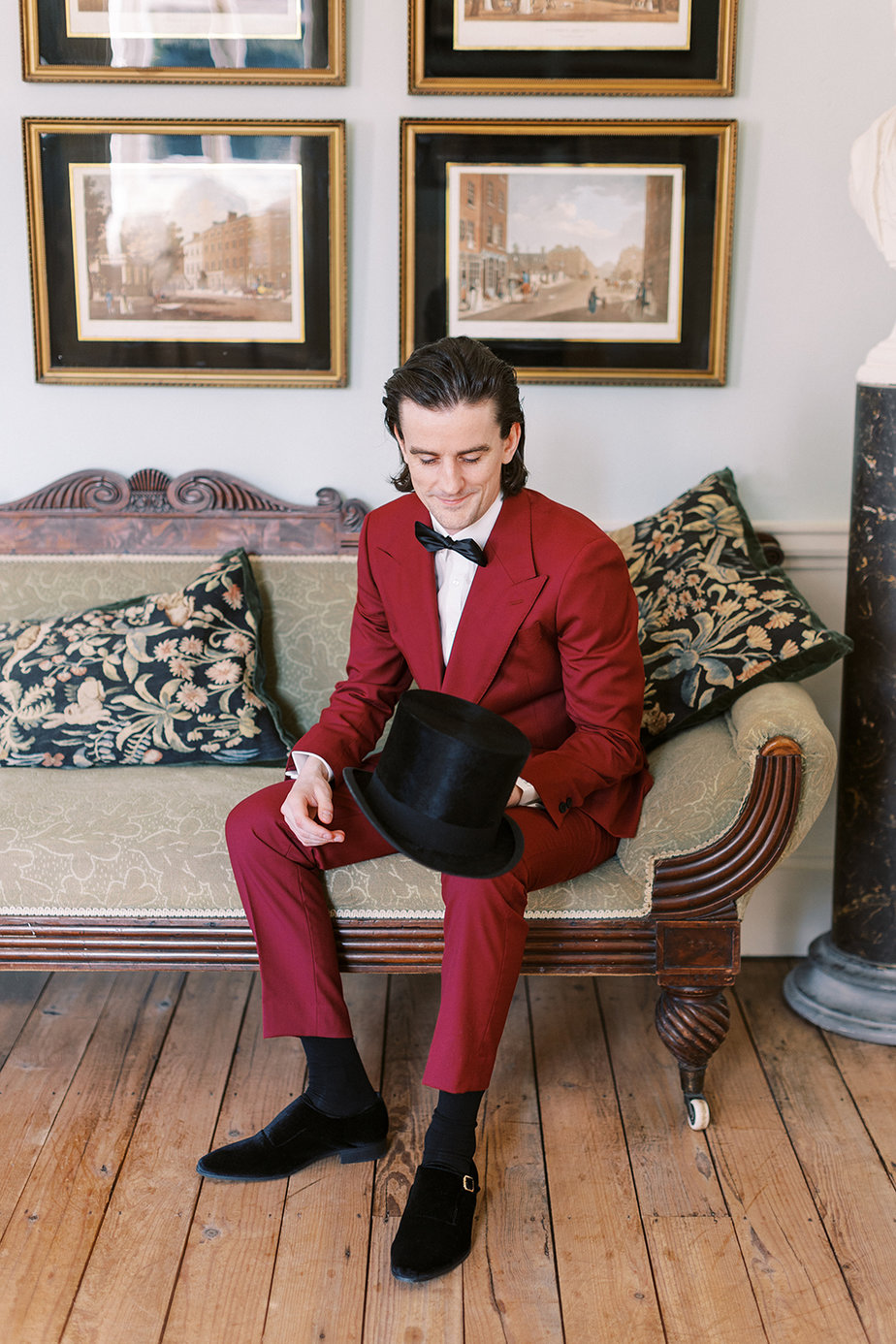 Groom wearing red suit interior manor house sitting holding hat