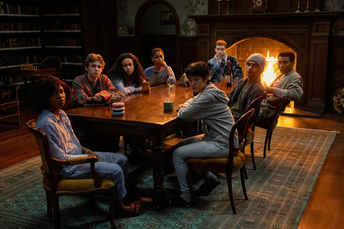 Adia as Cheri Ian, Igby Rigney as Kevin, Annarah Cymone as Sandra, Iman Benson as Ilonka, Aya Furukawa as Natsuki, Ruth Codd as Anya, Sauriyan Sapkota as Amesh, Chris Sumpter as Spencer all sitting around a table, in a library, next to a fireplace