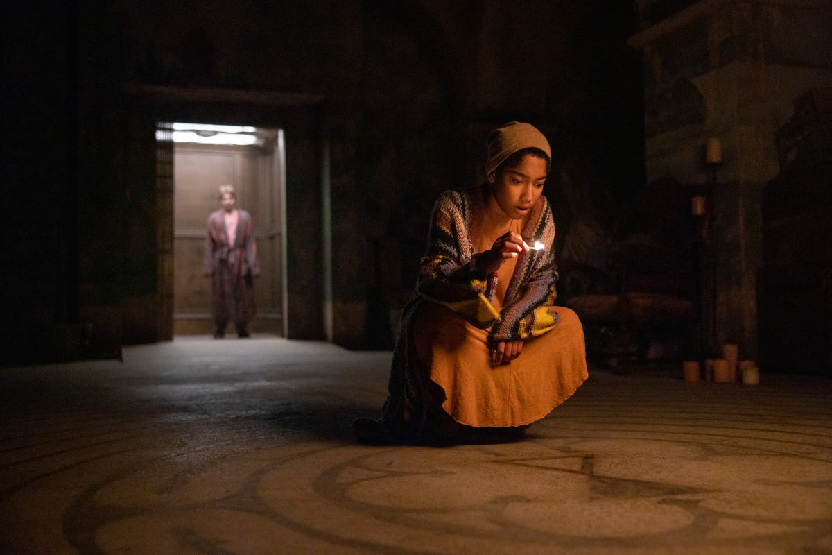Igby Rigney as Kevin standing in an elevator behind Iman Benson as Ilonka, who's holding a lit match and crouching down over strange markings on a basement floor