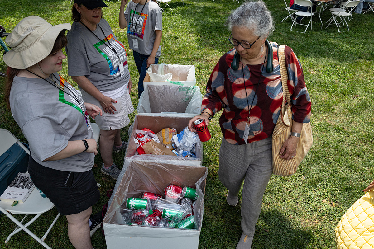 Three Festival volunteers in matching gray shirts and lanyards help a woman throw a Coca Cola can into the right disposing bin. Between the woman and volunteers are four cardboard bins marked Recycle, Landfill, and Compost.