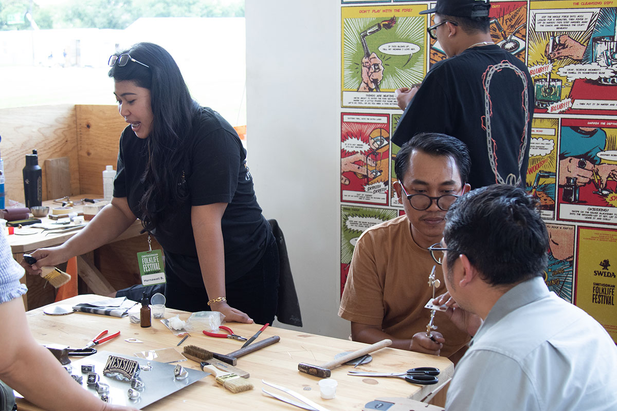 Around a work table set with metalworking tools, people chat.