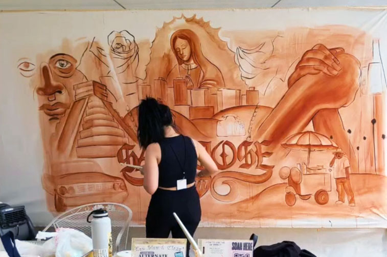 Facing away, a woman works on a sepia-colored mural wall depicting the Virgin Mary, a pyramid-shaped building, a street vendors, two clasped hands, and the words San Jose.