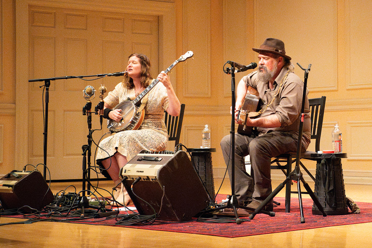 Two seated people perform on a warmly lit stage: a woman on banjo and a man on acoustic guitar, both singing.