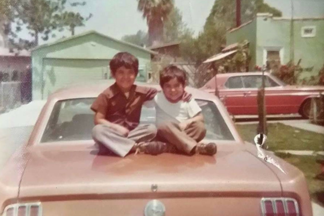 Two young boys sit cross-legged, arms around each other, on the hood of a classic car parked in a driveway. Old color film photo.