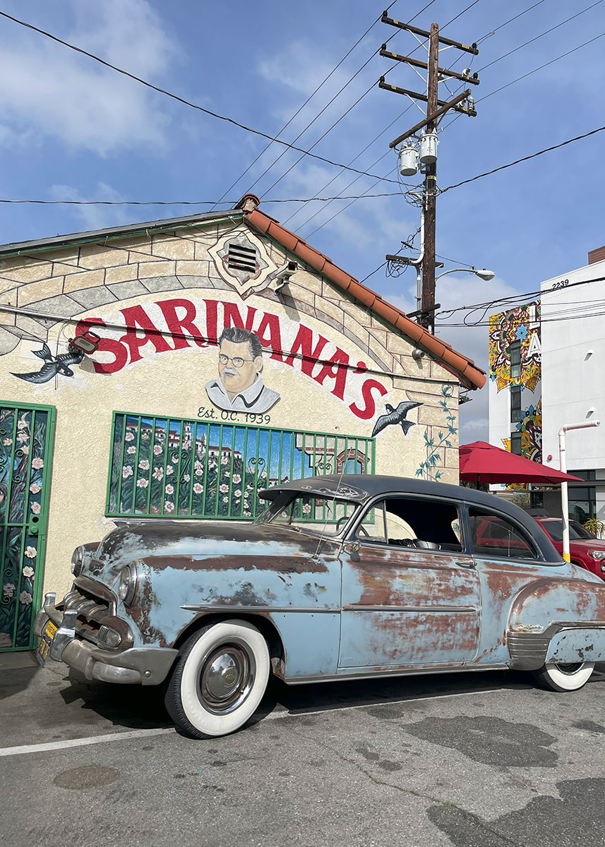 A pale blue classic car with rust spots parked on the street in front of a building.