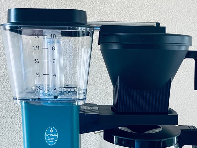 Front view of Moccamaster KBG.V. Select Coffee Maker in blue, a raised clear container connected to a spout for coffee grounds and a clear coffee pot with a handle.
