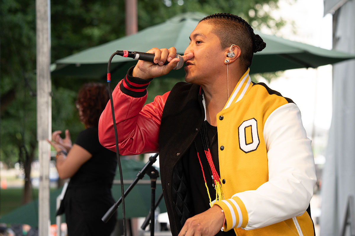 A man with a white, yellow, red, and black track jacket, an earpiece, and dark hair in cornrows raps into a microphone on stage. An ASL interpreter works off to the side.