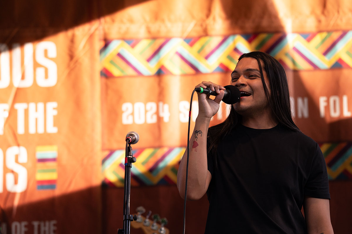A man with long, straight, dark hair sings into a microphone on stage.