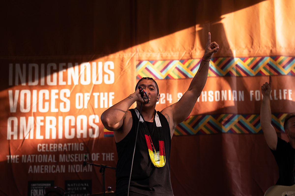 A man in a black tank top and yellow, red, black, and white beaded necklace raps on stage and raises one hand, pointing his index finger upward.