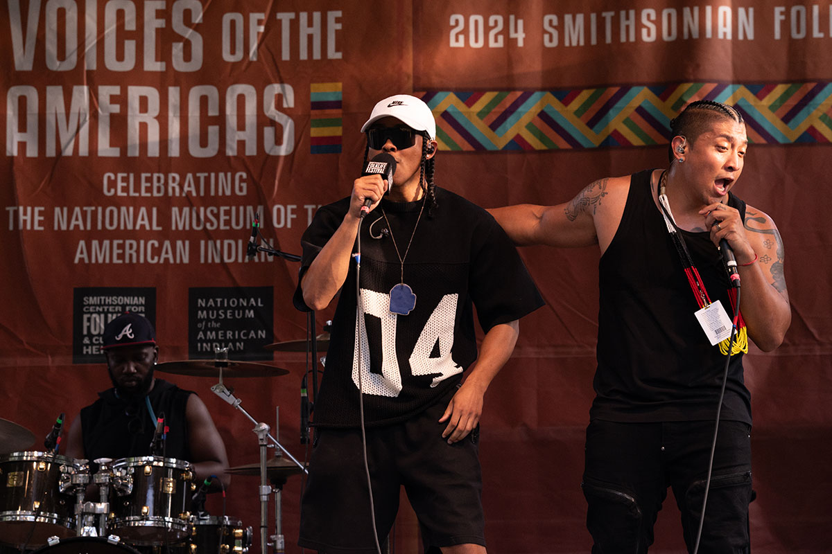 Two men in black clothing rap on stage, with a drummer on a drum kit playing in the background.