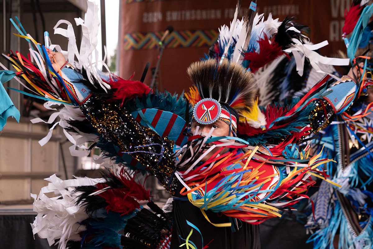 A  dancer performs in front of a stage, with colorful blue and red regalia and ribbons aloft with movement.