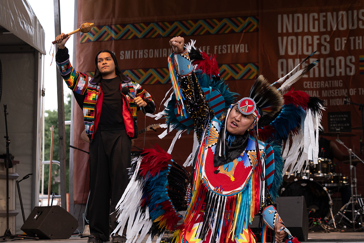 Two performers, one in black clothes with a rainbow quilted jacket and the other in full Native regalia, each raise a fist.