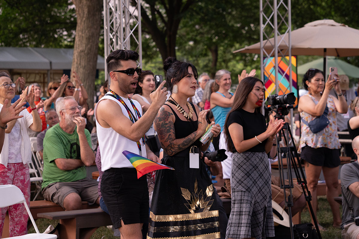 Audience members applaud, sitting and standing in a grassy area under trees and umbrellas.