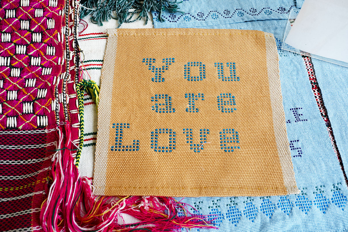 On a tabletop display of woven textile, a yellow square with the words YOU ARE LOVE woven in blue.
