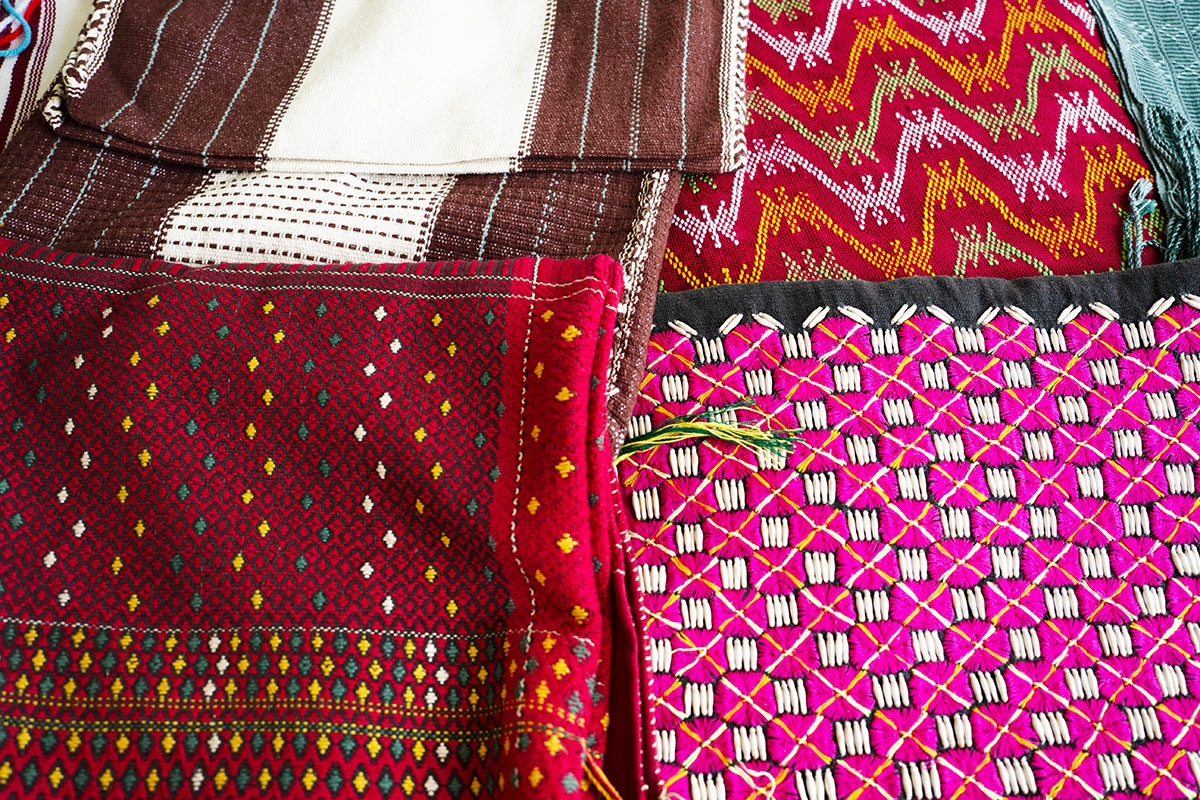Four woven textile arranged on a table: brown and white stripes; red with yellow, white, and green wavy stripes; red with a black diamond pattern and yellow, white, and green accents; and pink checkerboard pattern on black with yellow diagonal stripes and white threads or beads filling in the gaps between pink squares.