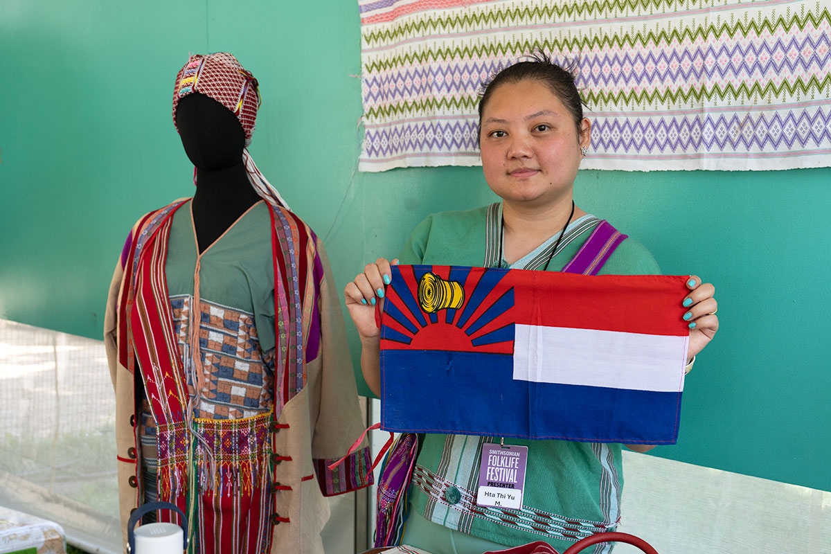 A young woman poses next to a mannequin; they both wear aqua-colored woven tops with colorful accents. She holds up a small flag, with red, white, and blue stripes on the right side, and a rising red sun under a yellow drum in the upper left corner.