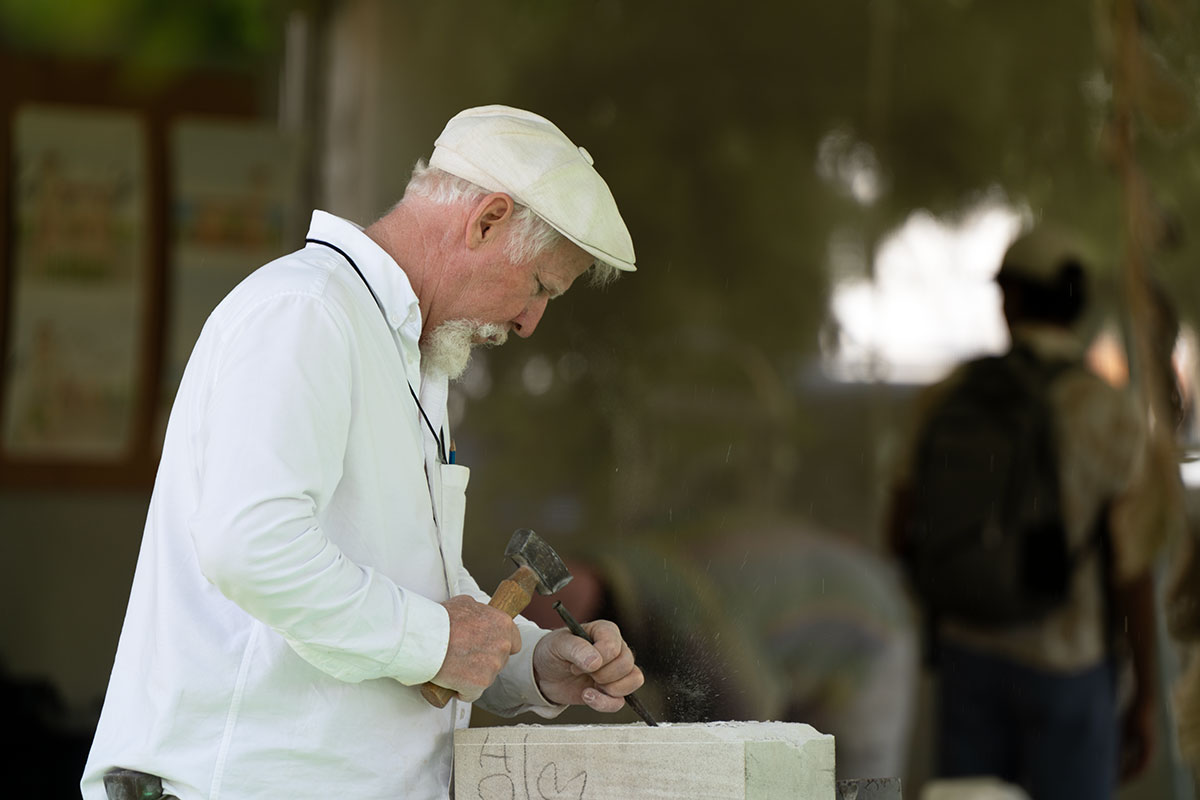A man with white hair and goatee uses hammer and chisel to carve into a gray stone block.
