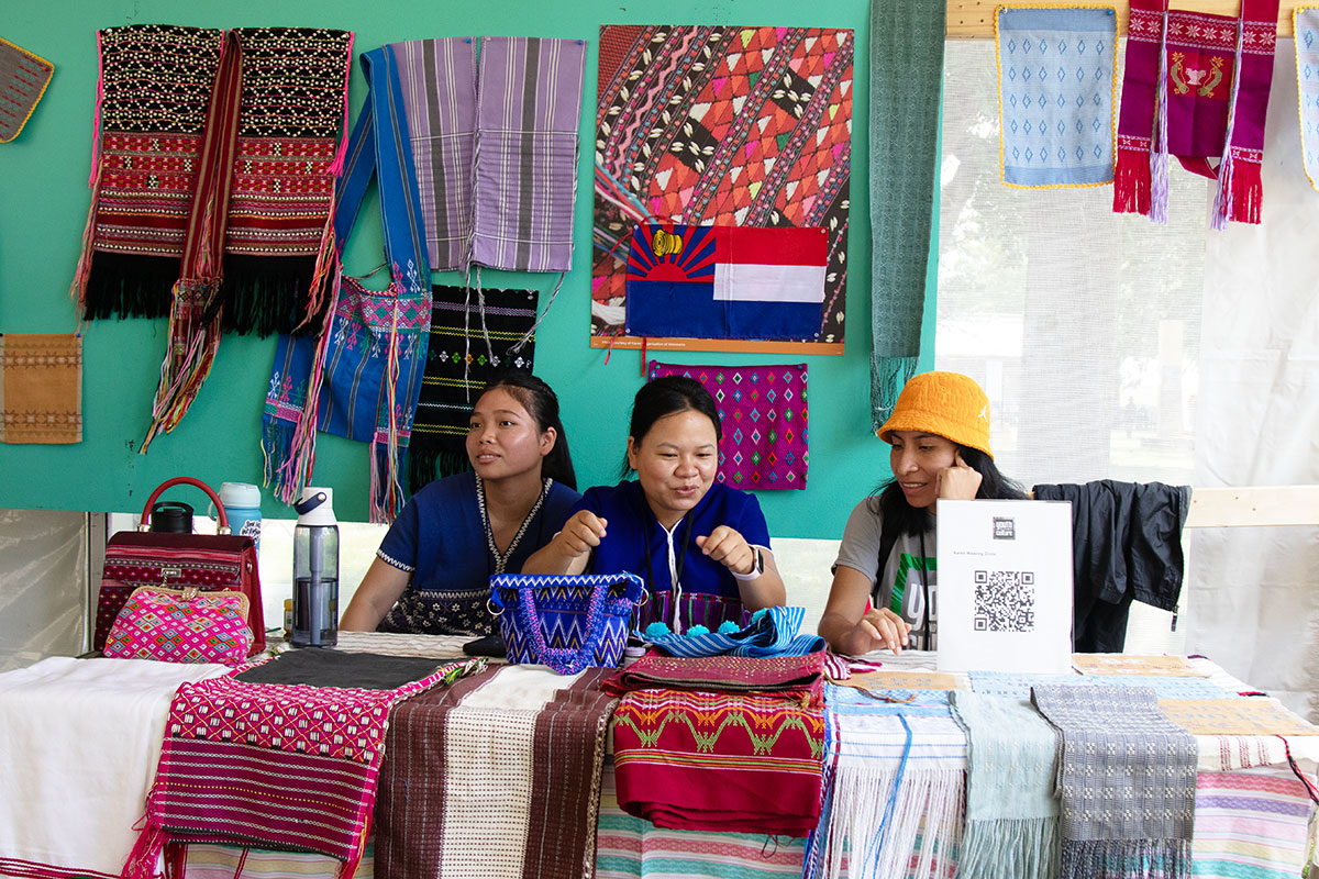 Three people sit behind a table set with various patterned woven textiles. The wall behind them is also festooned with various garments and cloths.
