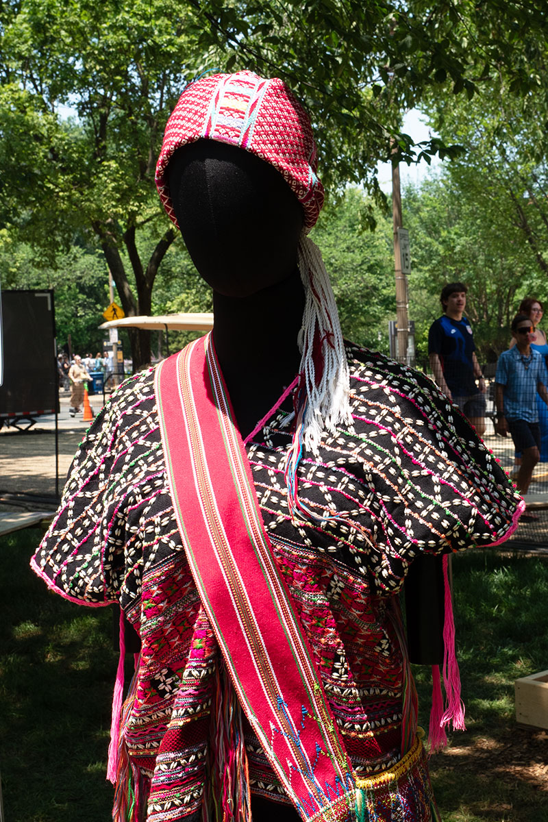 Outdoors, a mannequin is displayed wearing red and black woven headscarg, top, and sash.