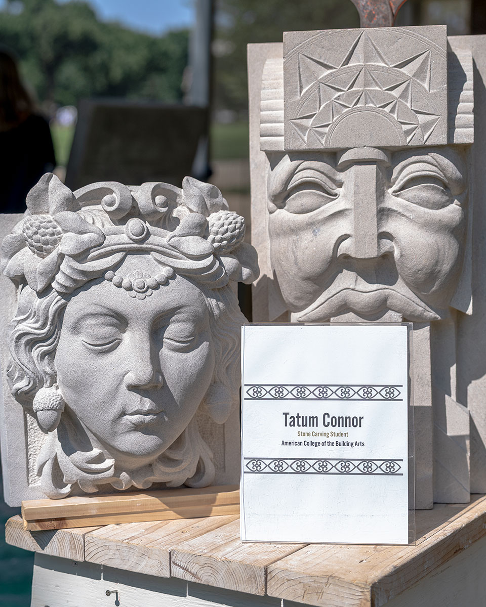 Two three-dimensional ornate stone sculptures depicting faecs with eyes closed. A tabletop sign in front of them reads the maker's name: Tatum Connor.