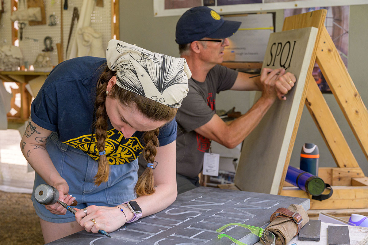 A young woman hunches over a slab of stone, using a hammer and chisel to carve letters reading Smithsonian Institution. Behind her, an older man also worked on a stone slab, painting the letters SPQR with his right hand, balanced on his left.