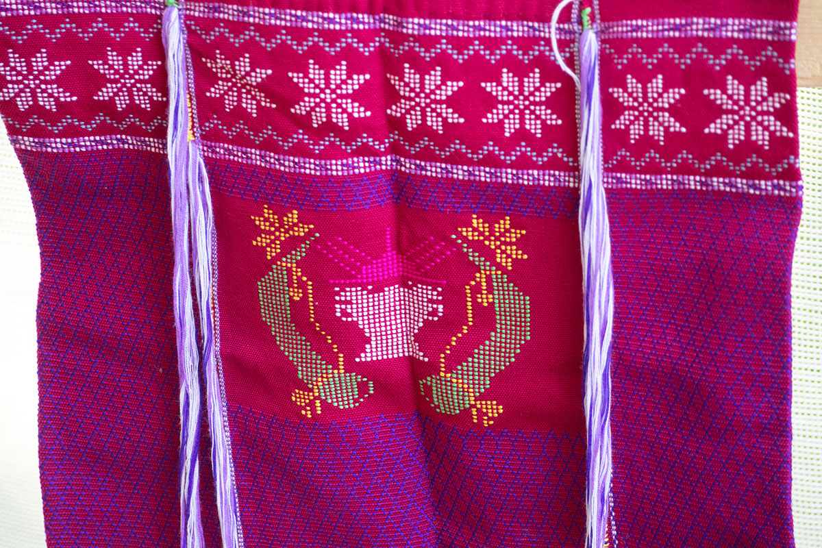 A hanging woven textile in red with purple diamond pattern. Across the top, white snowflake-like pattern with gray zigzag border. In the center, a pink rising sun flanked by two green animal horn shapes and a white drum shape below.
