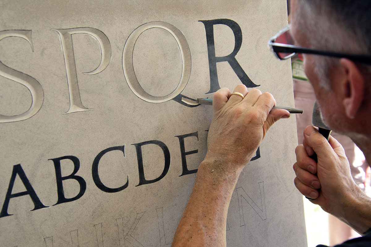 Close-up of the man carving the tail of a letter Q. The letters are painted black before carved out.