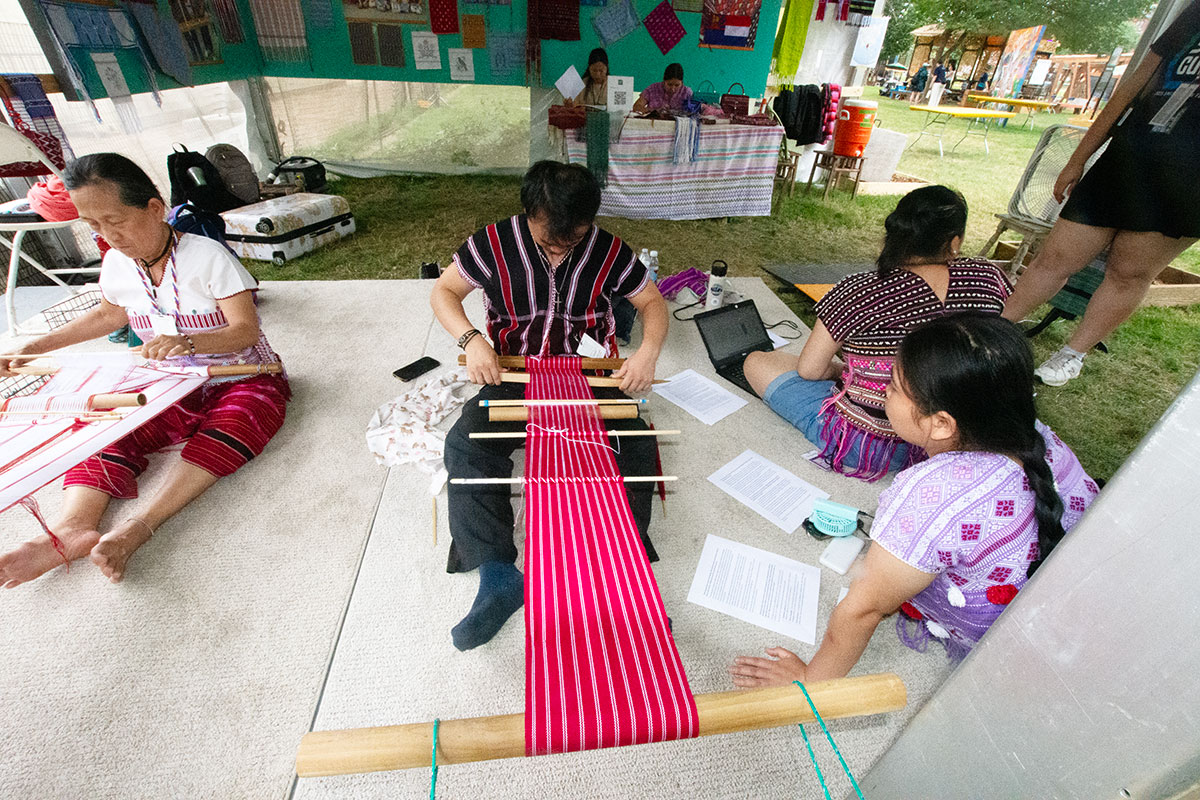 From a high angle, a man works on a red and white striped textile on a backstrap loom, seated on a low platform in the grass. An elder woman weaves to one side of him, and a younger woman works on a laptop  on the other side.