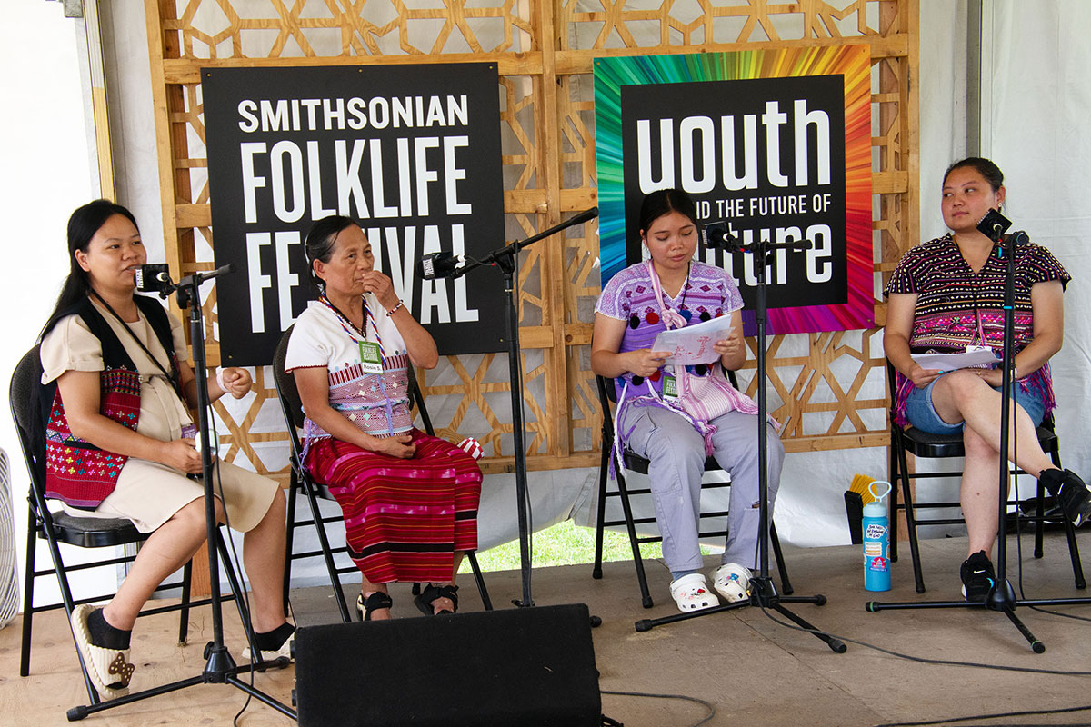 Four women sit onstage in front of microphones, all wearing woven tops. Two of the younger women wear Western pants and shorts, and sandals or tennis shoes.