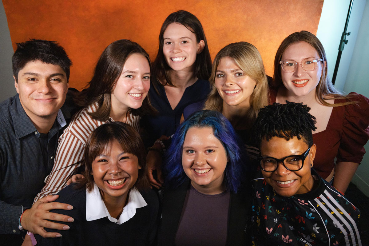 Eight people pose arm in arm in front of a painted orange background, all smiling.