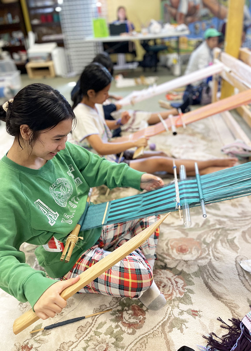 Indoors, three young girls sit on a floral rug, using backstrap looms attached to a wooden crossbar between pillars. The girl in front laughs as she weaves.