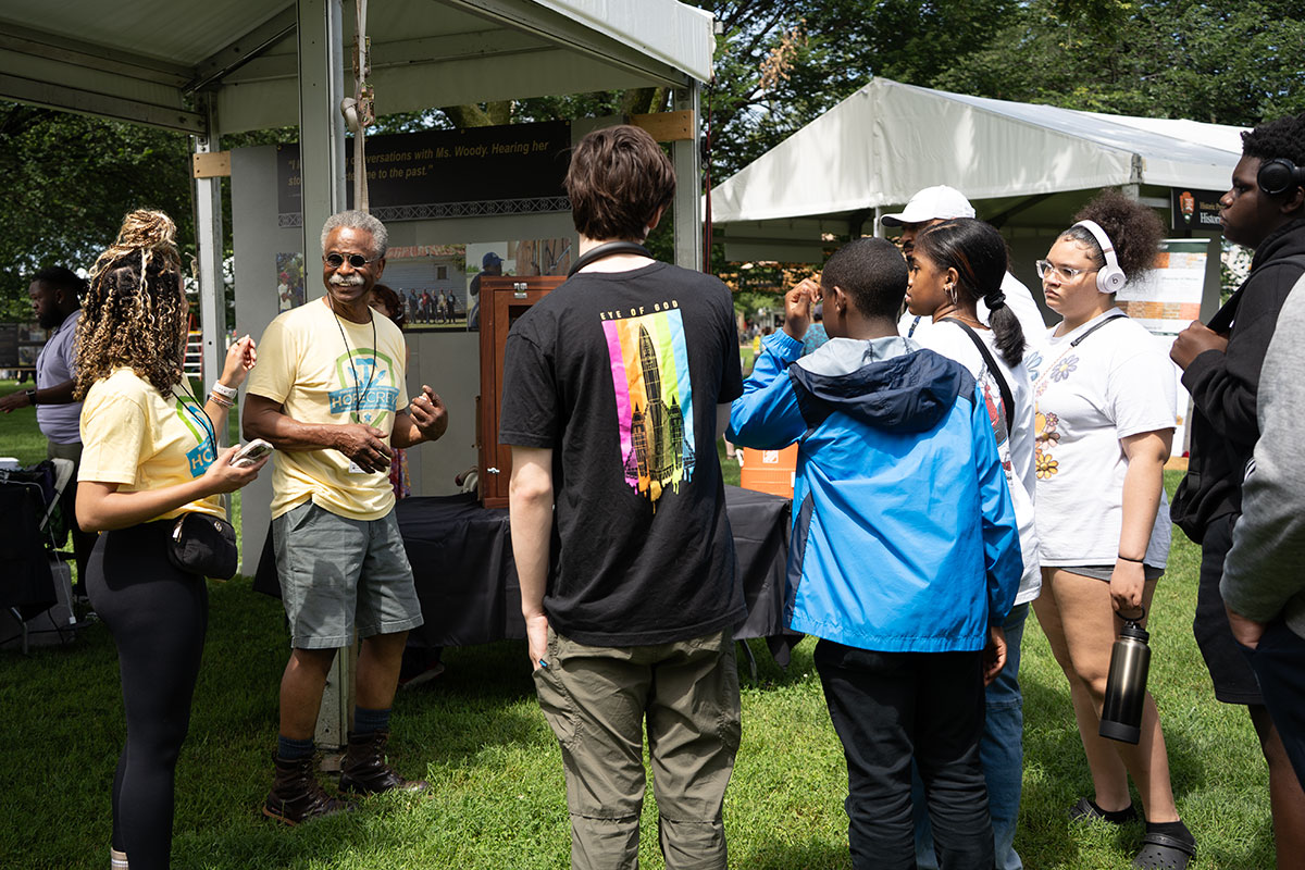 Outdoors among festival tents and trees, two people in yellow HOPE Crew shirts speak with a group of young visitors.