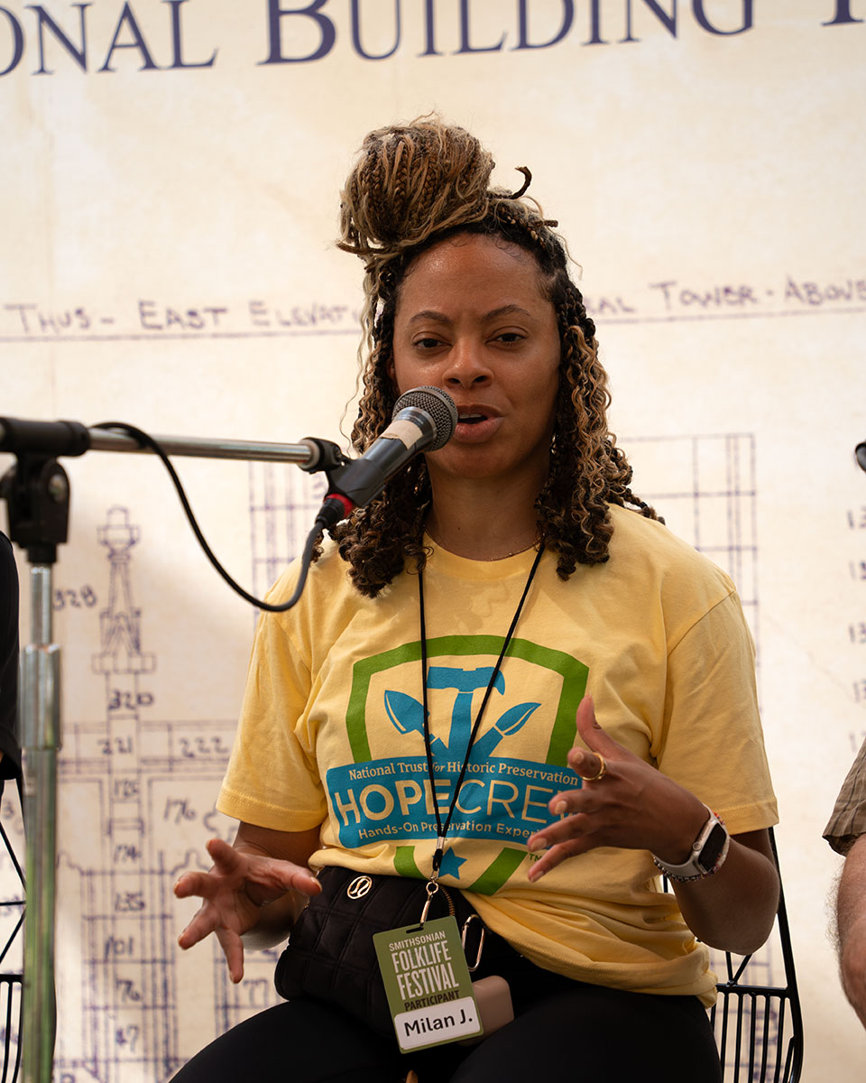 A woman in a yellow Hope Crew T-shirt speaks into a microphone, seated on stage.