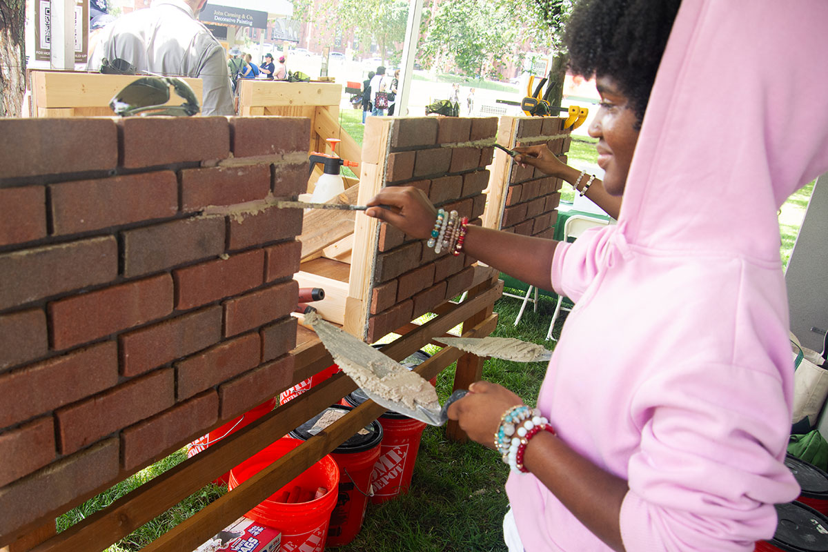 A young person in a pink hoodie smiles as they spread pale brown grout in between bricks on a demonstration wall. 