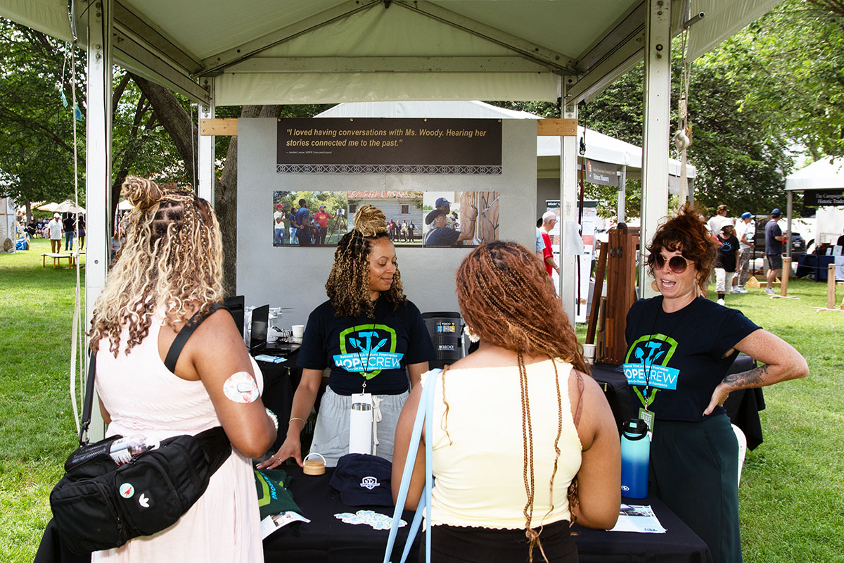 Four people stand around a table under a festival tent outdoors. Two of them wear matching black HOPE Crew T-shirts.