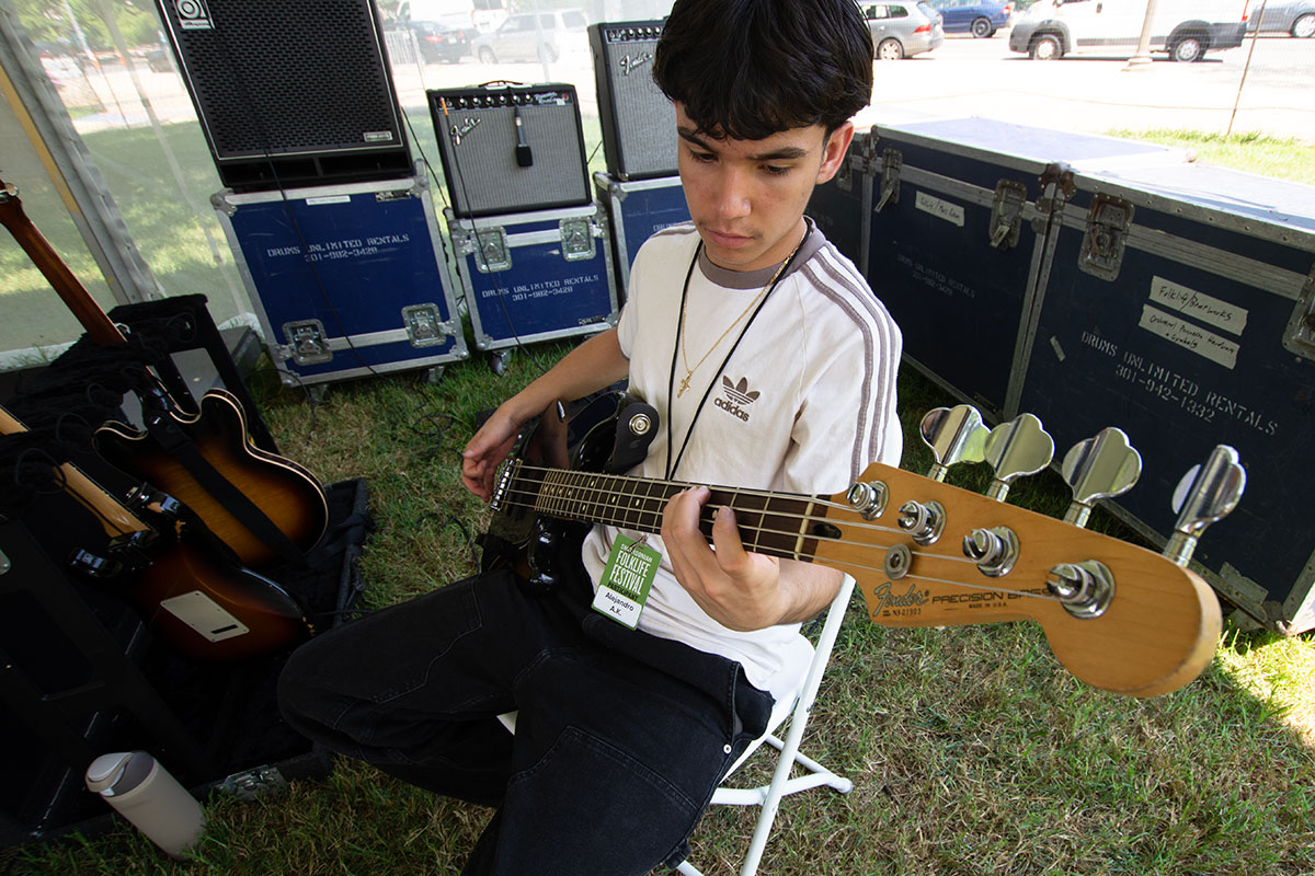 A teen boy wearing a white Adidas shirt sits under a tent surrounded by electric guitars and gear cases, playing an electric bass.