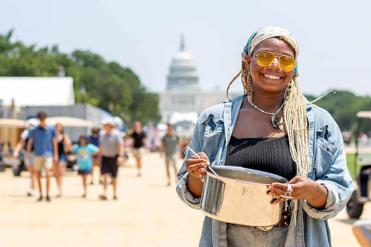 A young woman with yellow-tinted sunglasses and a bandana over her bleached braids holds a pot of soup with a ladle on the National Mall, with the U.S. Capitol Building in the background.