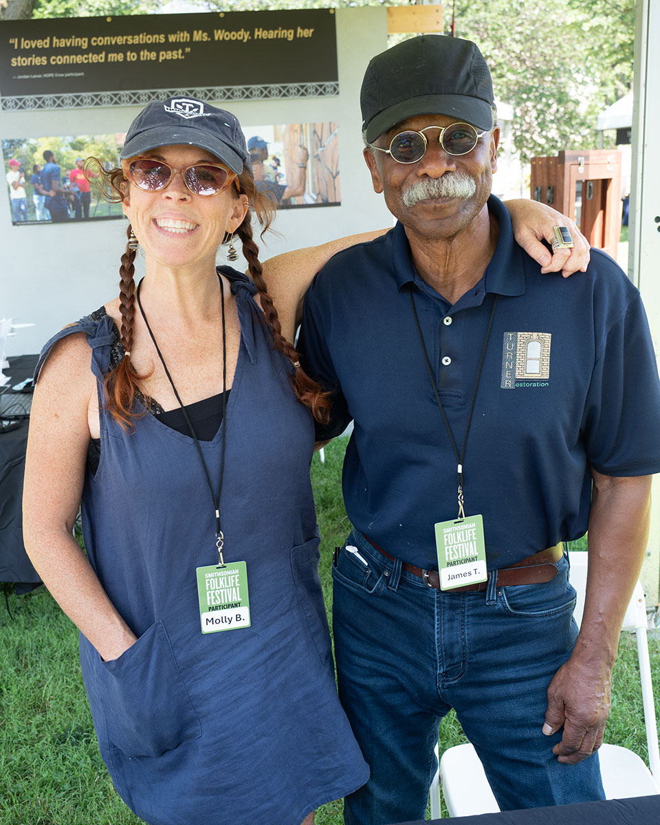 A woman in a blue apron with two long braids and a man with wire-rimmed sunglasses and a gray mustache pose arm in arm.