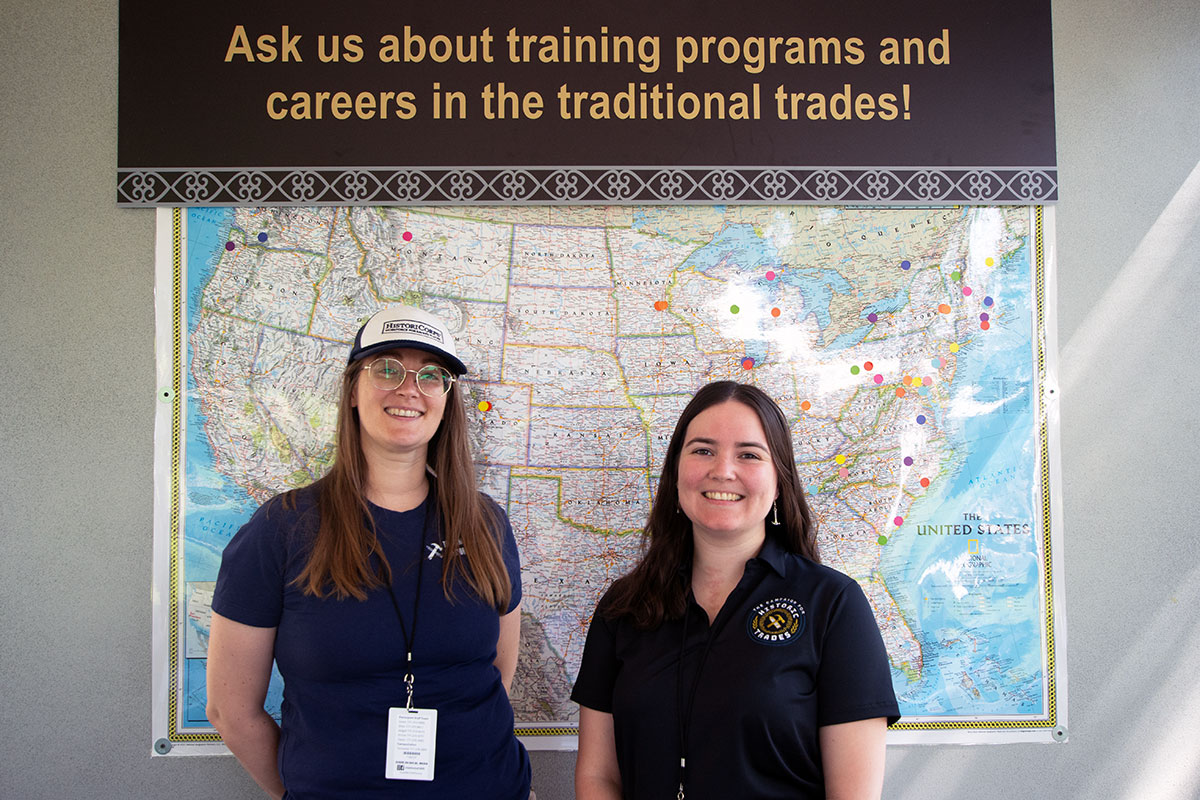 Two people stand and pose in front of a U.S. map with colorful dot markers across the country, especially concentrated in the Northeast. Above them, a sign reads Ask us about training programs and careers in the traditional trades!