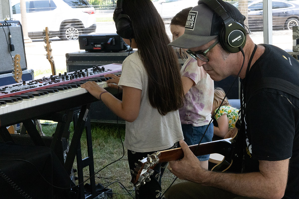 A man wearing a Folkways hat and headphones sits, concentrating as he plays electric guitar. Two young kids behind play an electric keyboard.