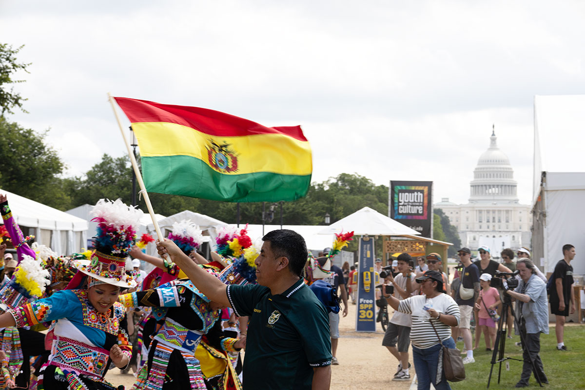 A group of dancers in colorful embroidered outfits and feathered hats dance on a walkway of the National Mall, as a crowd watches and takes photos and a man in the foreground waves a Bolivian flag, with red, yellow, and green stripes. In the distance is the U.S. Capitol Building.
